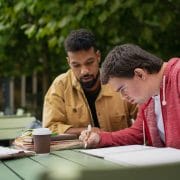 Young man with Down syndrome with his mentoring friend sitting outdoors in cafe and studying